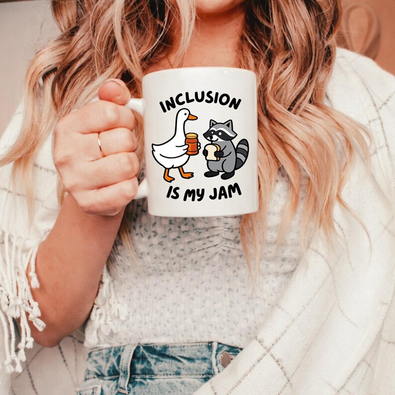 May include: White ceramic mug with the text "INCLUSION IS MY JAM". The mug features cartoon illustrations of a goose holding a jar of jam and a raccoon holding a piece of bread. The mug has a white handle.