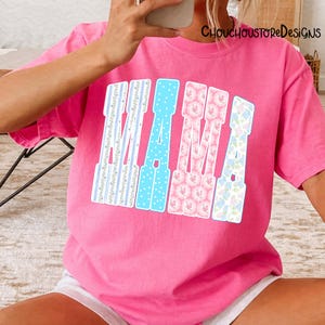 May include: A bright pink t-shirt with the word "MAMA" in a decorative font. The letters are in blue, pink, and white with floral and polka dot patterns. The shirt is a casual style.