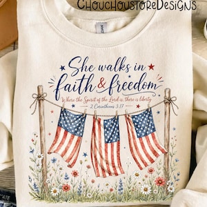 May include: Cream-colored sweatshirt with the text "She walks in faith & freedom" and American flag imagery. The design includes floral accents and the Bible verse 2 Corinthians 3:17.