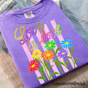 K&ouml;nnte beinhalten: Ein lilafarbenes T-Shirt mit dem Schriftzug "Love is Love" in Regenbogenfarben, zusammen mit bunten Blumen und rosa Streifen. Das T-Shirt ist aus weichem Stoff und eignet sich f&uuml;r den t&auml;glichen Gebrauch.