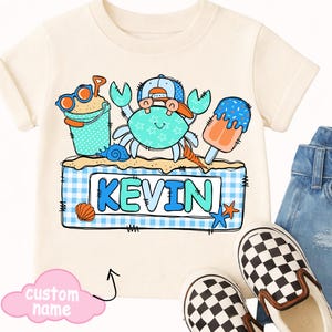 May include: Cream-colored t-shirt featuring a crab wearing a hat and sunglasses, with a sand bucket and popsicle. The name "KEVIN" is displayed on a blue and white checkered background. A pair of checkered slip-on shoes and denim jeans are also visible.