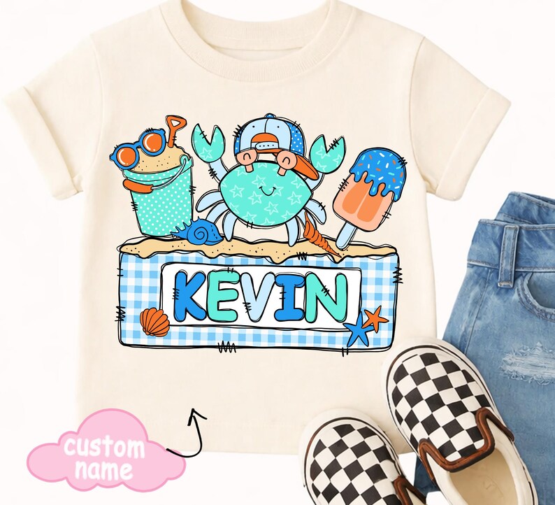 May include: Cream-colored t-shirt featuring a crab wearing a hat and sunglasses, with a sand bucket and popsicle. The name "KEVIN" is displayed on a blue and white checkered background. A pair of checkered slip-on shoes and denim jeans are also visible.