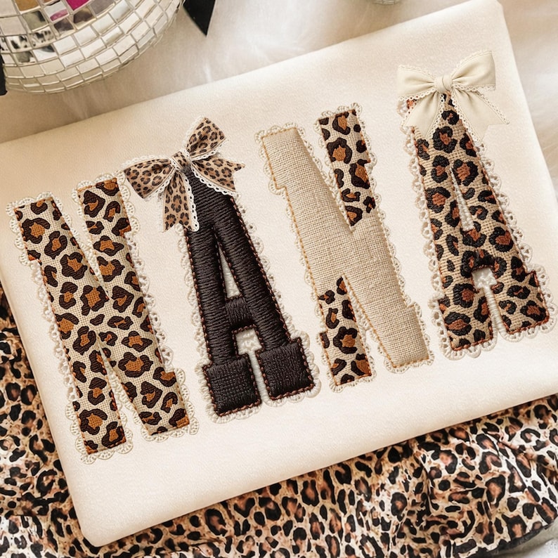 May include: Cream-colored fabric with the word "NANA" embroidered in a mix of leopard print and solid brown. The letters are outlined with lace and adorned with small bows. The fabric is resting on a leopard print surface.