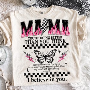 May include: Cream-colored t-shirt with the word "MAMA" in black, outlined in pink flames. The shirt features a butterfly graphic and the text "YOU'RE DOING BETTER THAN YOU THINK" and "I believe in you."