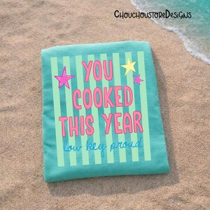 May include: A folded turquoise beach towel with vertical light green stripes. The towel features the text "YOU COOKED THIS YEAR" in pink, with a yellow star and pink star accents. The phrase "low key proud" is written below.