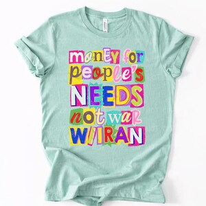 May include: A light teal t-shirt with the text "money for people's NEEDS not war W/IRAN" in colorful, cut-out style letters. The text is arranged in a stacked format, with each word or phrase on a separate line.