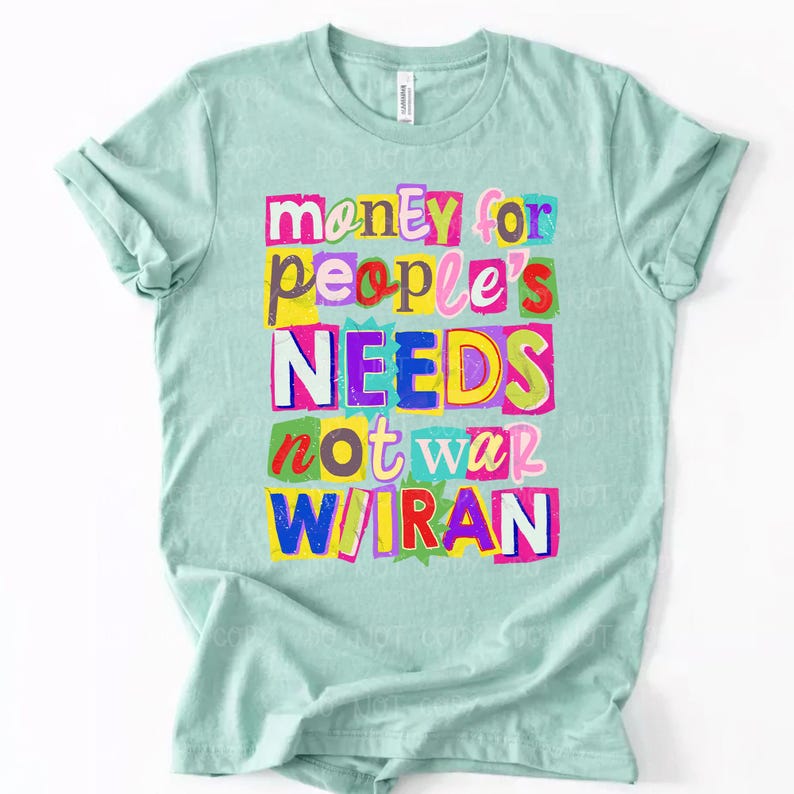 May include: A light teal t-shirt with the text "money for people's NEEDS not war W/IRAN" in colorful, cut-out style letters. The text is arranged in a stacked format, with each word or phrase on a separate line.