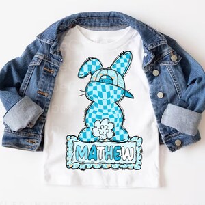 May include: White t-shirt featuring a blue and white checkered bunny wearing a cap. The bunny design includes a flower and the name "MATHEW" in a blue frame. A denim jacket is draped over the shirt.