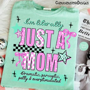 May include: A light green t-shirt with the text "I'm literally JUST A MOM" in pink and black checkered letters. The shirt has star and bow graphics. The text "Dramatic, sarcastic, petty & overstimulated" is also on the shirt.