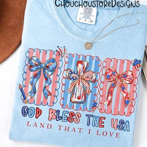 May include: Light blue t-shirt with a patriotic design. The shirt features three framed images of bows and a cross, all decorated with red, white, and blue patterns. The text "GOD BLESS THE USA LAND THAT I LOVE" is printed below the images.