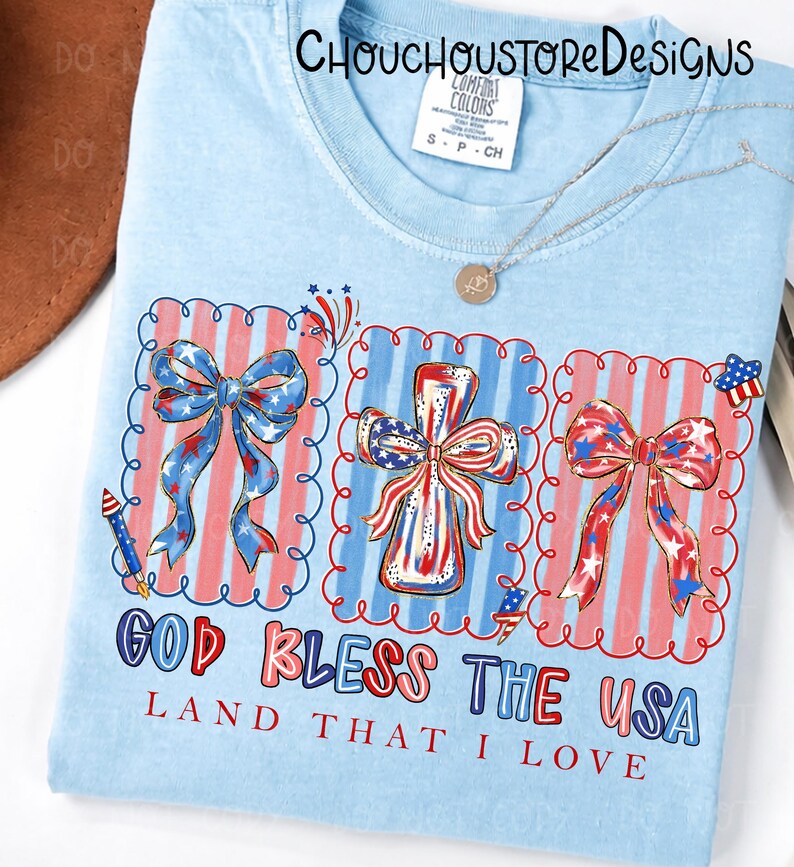 May include: Light blue t-shirt with a patriotic design. The shirt features three framed images of bows and a cross, all decorated with red, white, and blue patterns. The text "GOD BLESS THE USA LAND THAT I LOVE" is printed below the images.