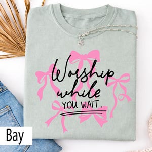 May include: A light green t-shirt with the words "Worship while you wait" in black script and pink bows. A silver necklace with a pendant is draped across the shirt. The shirt is folded on top of blue jeans.