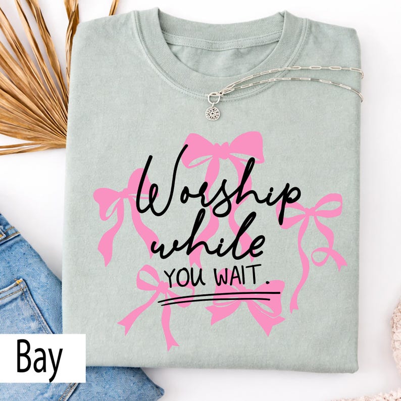 May include: A light green t-shirt with the words "Worship while you wait" in black script and pink bows. A silver necklace with a pendant is draped across the shirt. The shirt is folded on top of blue jeans.