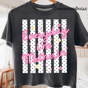May include: Dark grey t-shirt with the text "Everyday I'm Motherin" in pink script. The design features white vertical stripes with black polka dots, and small pink and yellow flower graphics.