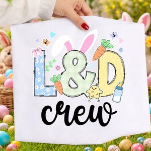 May include: White Easter-themed graphic tee with the words "L & D crew" in black script. The design includes bunny ears, carrots, a chick, and a baby bottle. The letters "L" and "D" are decorated with floral and polka dot patterns, and the ampersand is green.