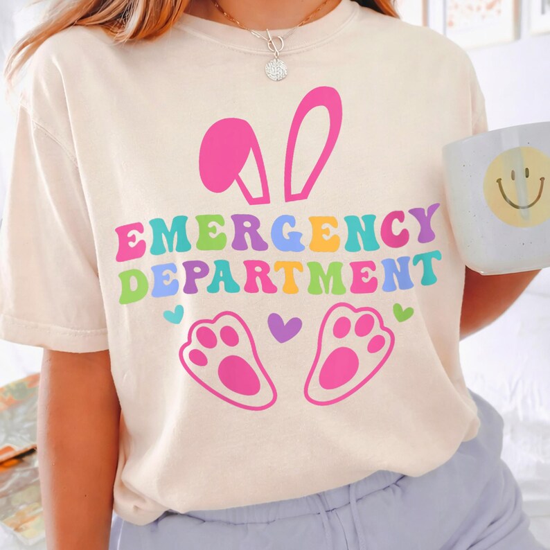 May include: Cream-colored t-shirt with the words "EMERGENCY DEPARTMENT" in colorful, bubble letters. Above the text are pink bunny ears, and below are pink paw prints. A white mug with a yellow smiley face is held in the hand.
