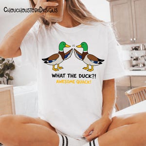 May include: White t-shirt featuring a cartoon design of two ducks with green heads, brown bodies, and yellow feet. The text "WHAT THE DUCK?! AWESOME QUACK!" is printed below the ducks. The brand name "ChouchoustoreDesigns" is at the top.