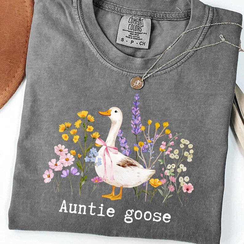 May include: Gray t-shirt with a floral design and a goose wearing a pink bow. The text "Auntie goose" is printed at the bottom. A silver necklace with a pendant is on the shirt.