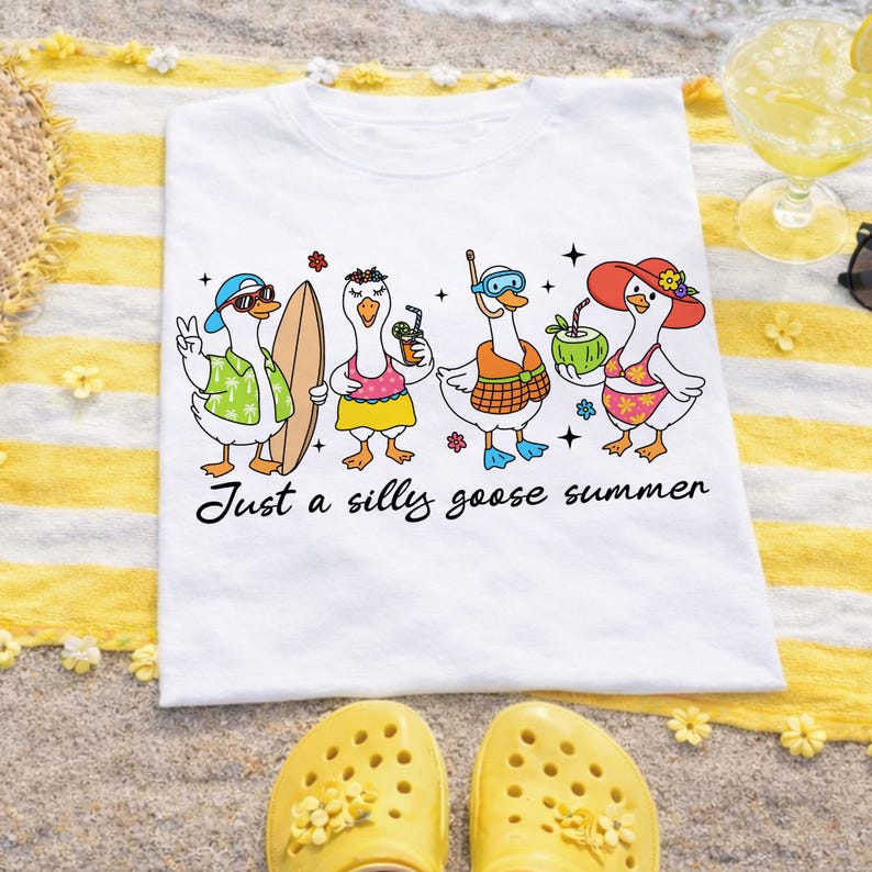 May include: White t-shirt featuring a cartoon illustration of four geese enjoying summer activities. One goose wears sunglasses and a Hawaiian shirt, another holds a surfboard, and the others have drinks. The text "Just a silly goose summer" is below the illustration.