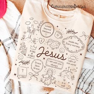 May include: Cream-colored sweatshirt with a black graphic design featuring the word "Jesus" and various religious symbols and phrases, including a cross, sheep, and "Light of the World."
