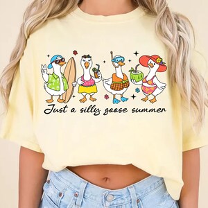 May include: A light yellow t-shirt featuring a whimsical illustration of four cartoon geese, each accessorized for summer fun. One goose wears sunglasses and a Hawaiian shirt, another holds a surfboard, and the others have drinks and beach attire. The text "Just a silly goose summer" is below.