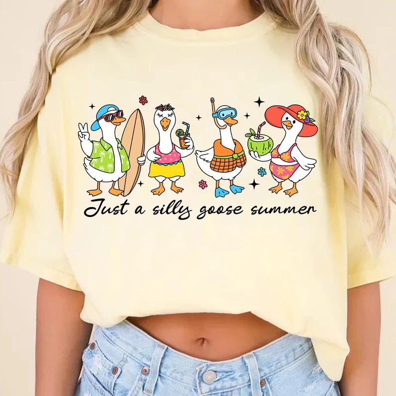 May include: A light yellow t-shirt featuring a whimsical illustration of four cartoon geese, each accessorized for summer fun. One goose wears sunglasses and a Hawaiian shirt, another holds a surfboard, and the others have drinks and beach attire. The text "Just a silly goose summer" is below.