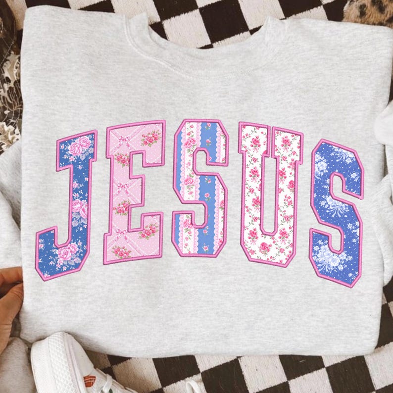 Puede incluir: Una sudadera gris claro con la palabra "JESUS" en letras grandes con estampado floral. Las letras est&aacute;n delineadas en rosa y presentan dise&ntilde;os florales azules y rosas, creando una est&eacute;tica vintage.