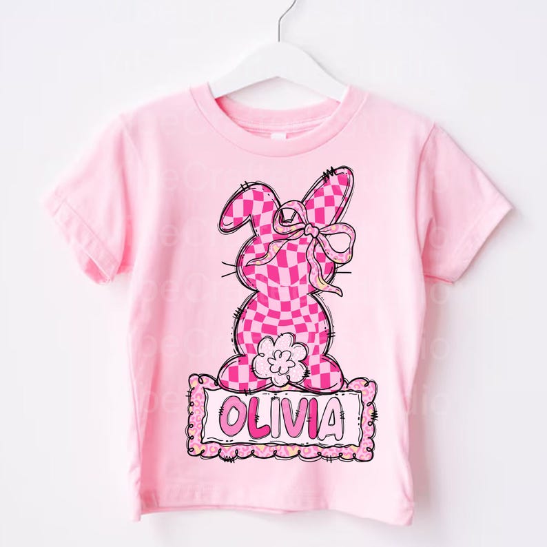 May include: A pink t-shirt featuring a cartoon bunny design with a checkered pattern in shades of pink. The bunny has a bow and is above a sign that says "OLIVIA" in pink letters. The shirt is on a white hanger.
