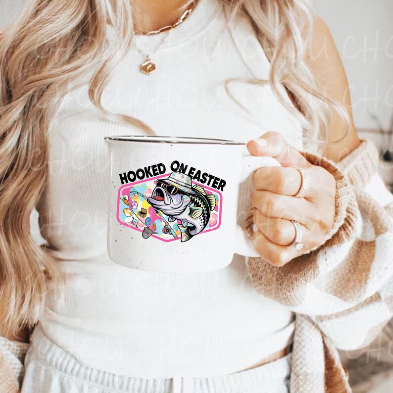 Pode incluir: Caneca de cer&acirc;mica branca com borda preta. A caneca apresenta um gr&aacute;fico colorido de um peixe usando &oacute;culos de sol e chap&eacute;u, com o texto "HOOKED ON EASTER" em preto. O fundo do gr&aacute;fico &eacute; um design rosa e azul.