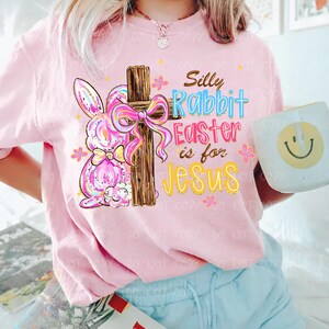 Peut inclure: T-shirt rose avec un motif color&eacute; repr&eacute;sentant un lapin, une croix en bois avec un n&oelig;ud et le texte "Silly Rabbit Easter is for Jesus". Le motif comprend des fleurs et une tasse smiley.