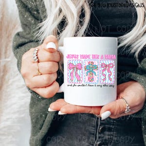 May include: White ceramic mug featuring the text "JESUS MADE HER A MAMA" in pink, with three decorative images of bows and a cross. The mug also includes the phrase "and she wouldn't have it any other way".