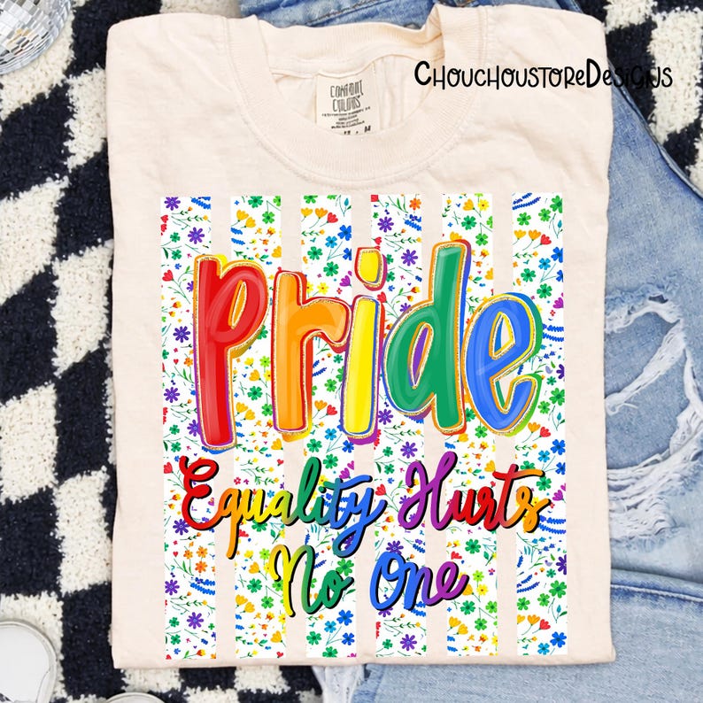May include: Cream-colored t-shirt with the word "Pride" in rainbow colors, and the phrase "Equality Hurts No One". The design is set against a floral and striped background. The shirt is from the brand "Comfort Colors".