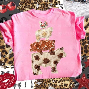 May include: A pink t-shirt featuring a stacked animal design. The design includes a cow, pig, sheep, and chicken, each with unique patterns and textures. The cow is brown and white, the pig is brown with floral accents, the sheep is cream with sunburst details, and the chicken has floral accents.
