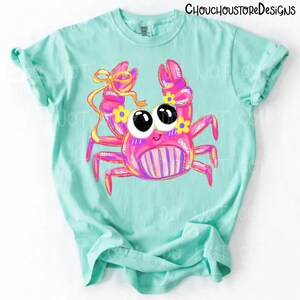 May include: A light blue t-shirt featuring a cartoon crab design. The crab is pink with yellow and purple accents, large black eyes, and a yellow bow. The text "ChouchoustoreDesigns" is at the top.