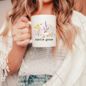 May include: White ceramic mug featuring a watercolor illustration of a goose surrounded by wildflowers. The mug has the text "Auntie goose" in black. The design includes yellow, purple, and pink flowers, and a pink bow.