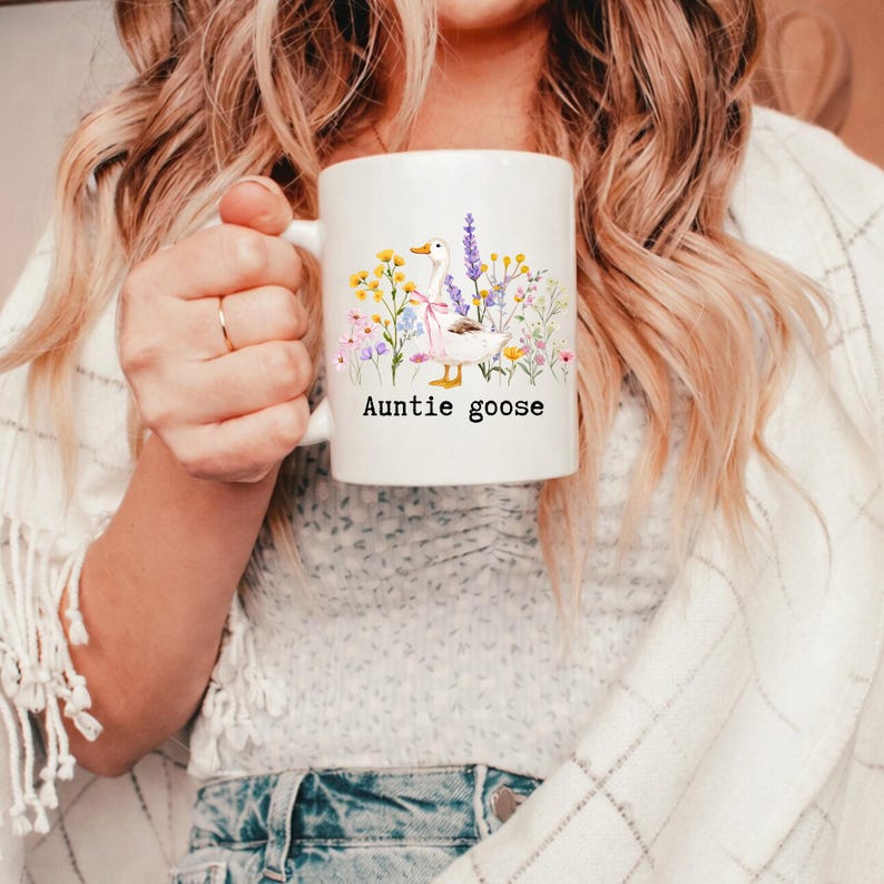 May include: White ceramic mug featuring a watercolor illustration of a goose surrounded by wildflowers. The mug has the text "Auntie goose" in black. The design includes yellow, purple, and pink flowers, and a pink bow.
