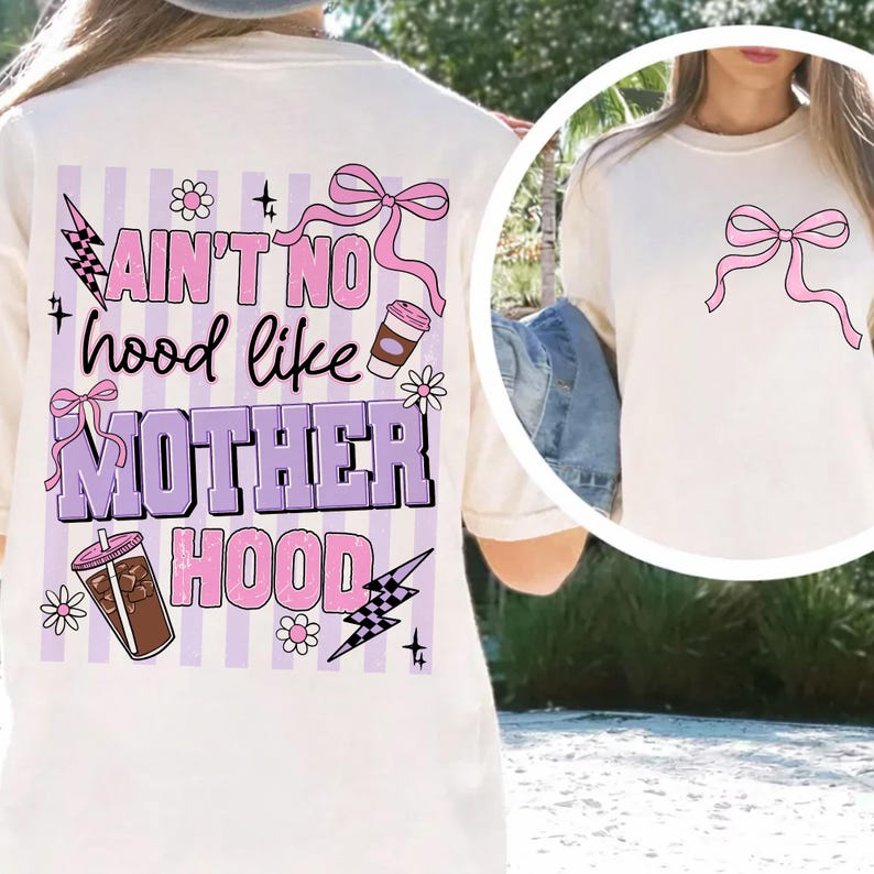 May include: Cream-colored t-shirt with a pink bow graphic on the front and a larger design on the back. The back design features the text "AIN'T NO hood like MOTHER HOOD" in pink and purple, along with coffee cups, flowers, and lightning bolts.
