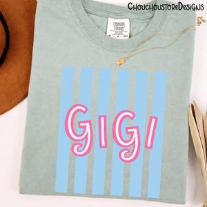 Puede incluir: Camiseta verde claro con el nombre "GIGI" en letras rosas y blancas, sobre un fondo de rayas verticales azul claro. La camiseta tiene cuello redondo y una peque&ntilde;a etiqueta en el cuello. Una cadena de oro est&aacute; drapeada sobre la camiseta.