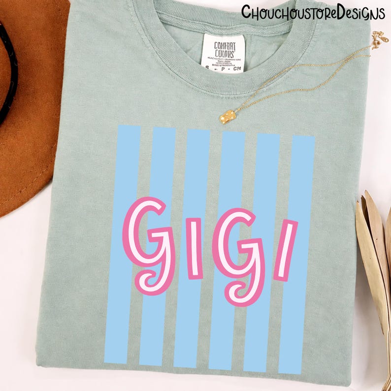 Puede incluir: Camiseta verde claro con el nombre "GIGI" en letras rosas y blancas, sobre un fondo de rayas verticales azul claro. La camiseta tiene cuello redondo y una peque&ntilde;a etiqueta en el cuello. Una cadena de oro est&aacute; drapeada sobre la camiseta.