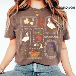 May include: A brown t-shirt with various framed illustrations. The illustrations include a teapot and teacup, strawberries, a duck with a bow, a basket of cherries, lavender, and a duck with a blue bow. The text "LOUCHOUSTOREDESIGNS" is visible on the shirt.