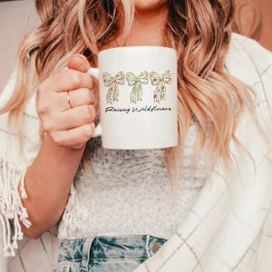 May include: White ceramic mug featuring three decorative bows with floral patterns in pink, green, and yellow. The mug has the text "Raising Wildflowers" in a handwritten style font.