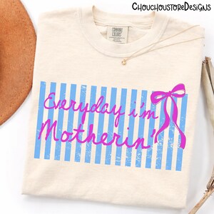 Pu&ograve; includere: T-shirt color crema con la scritta "Everyday I'm Motherin'" in rosa su sfondo a righe blu. Un fiocco rosa &egrave; sul lato destro. La maglietta ha un'etichetta Comfort Colors.