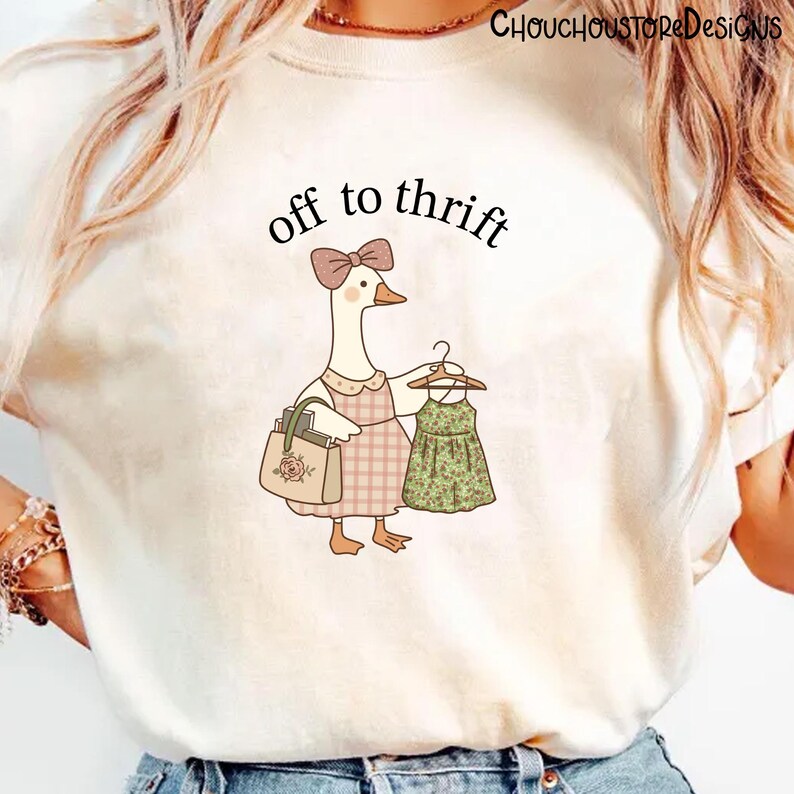 May include: A cream-coloured t-shirt with a cartoon goose wearing a pink bow, holding a dress and a purse. The goose wears a pink plaid apron and the text "off to thrift" is above the goose. The purse has a rose design.