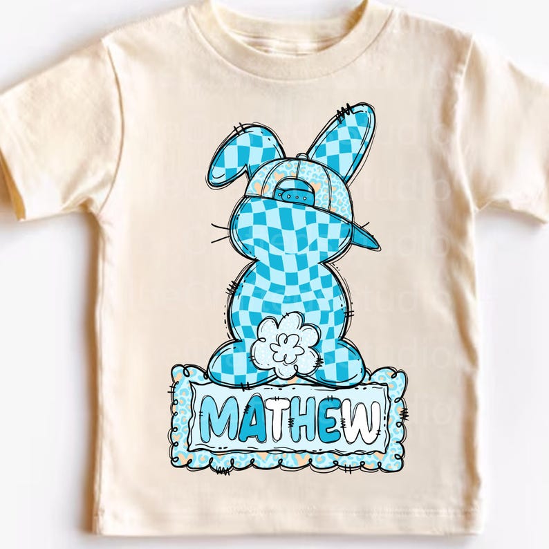 May include: A cream-colored toddler t-shirt featuring a cartoon bunny wearing a blue checkered hat. The bunny is also blue checkered. The name "MATHEW" is printed in blue on a decorative frame at the bottom of the shirt.
