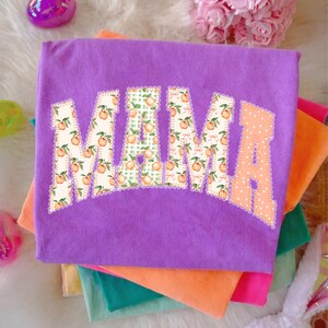 May include: A stack of colorful t-shirts, with a purple shirt on top. The purple shirt features the word "MAMA" in a decorative font, with peach and green patterns. The shirts are in shades of orange, pink, and green.