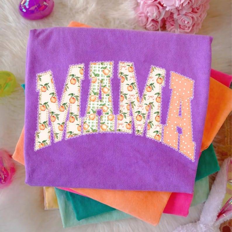 May include: A stack of colorful t-shirts, with a purple shirt on top. The purple shirt features the word "MAMA" in a decorative font, with peach and green patterns. The shirts are in shades of orange, pink, and green.