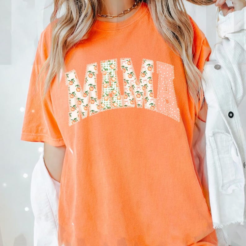 May include: Orange t-shirt with the word "MAMA" in a patchwork design. The letters are filled with peach and floral patterns. The shirt has short sleeves and a relaxed fit.