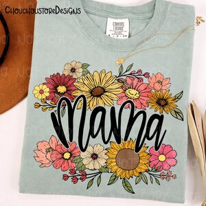 May include: A light green t-shirt with the word "Mama" in black script, surrounded by a colorful floral arrangement. The flowers include sunflowers, daisies, and other blooms in shades of yellow, pink, and red. The shirt has a Comfort Colors label.