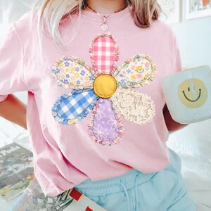 May include: A light pink t-shirt featuring a large, appliqued flower design. The flower has a yellow center and petals made from various patterned fabrics, including gingham, floral, and lace. A light blue mug with a yellow smiley face is held in the hand.