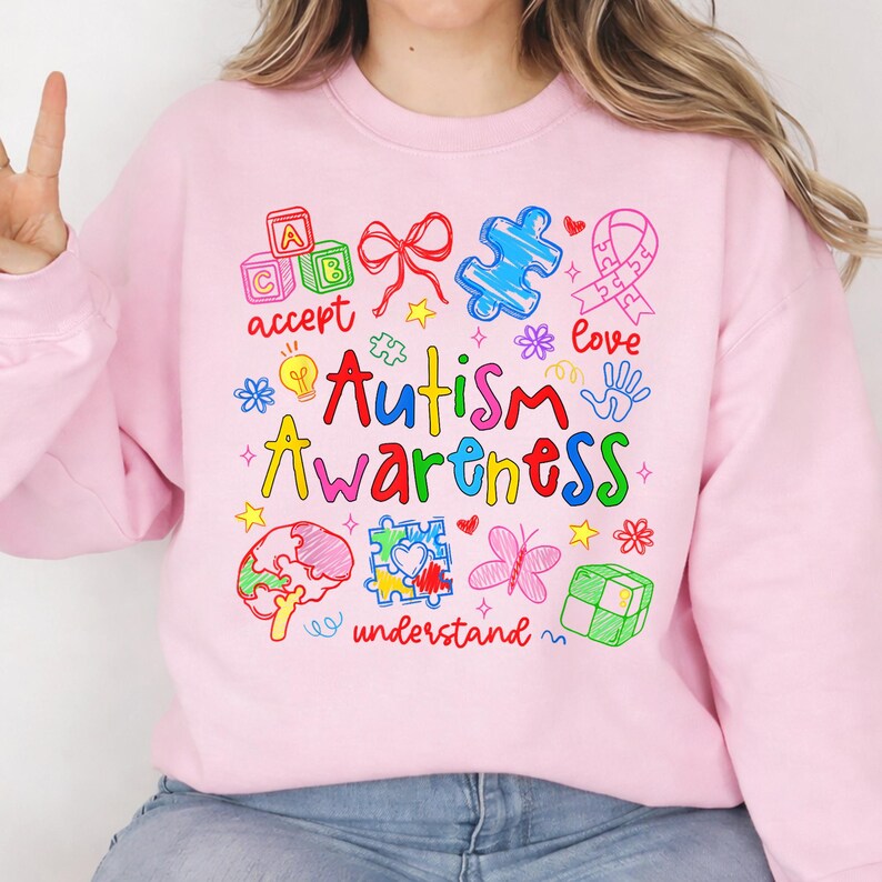 May include: Pink sweatshirt with a colorful graphic design promoting Autism Awareness. The design includes puzzle pieces, a ribbon, and the words "Autism Awareness" in a playful font, along with other related words and symbols.